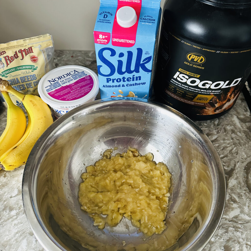 Mashed banana muffin batter in a metal mixing bowl, surrounded by ingredients including Silk Protein almond & cashew milk, PVL IsoGold whey protein, chia seeds, cottage cheese, and fresh bananas.