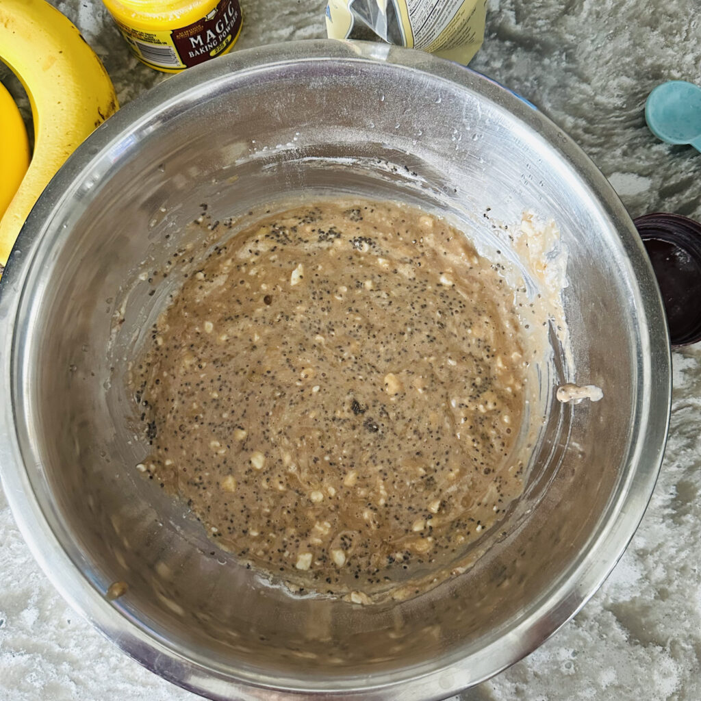 Mashed banana muffin batter in a metal mixing bowl, surrounded by ingredients including Silk Protein almond & cashew milk, PVL IsoGold whey protein, chia seeds, cottage cheese, and fresh bananas.
