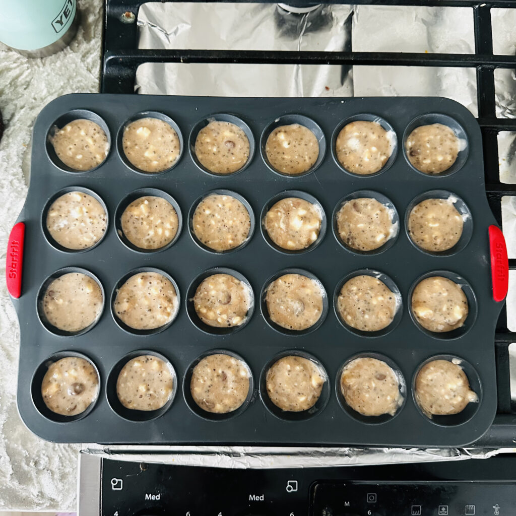 Mini muffin batter in a 24-cup silicone muffin tray, filled and ready to bake, placed on a stovetop with foil-lined surface below.