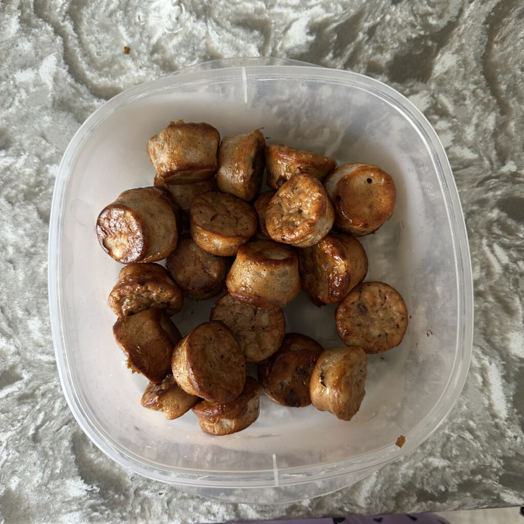 Cooked mini protein muffins stacked inside a clear plastic storage container on a marble countertop.