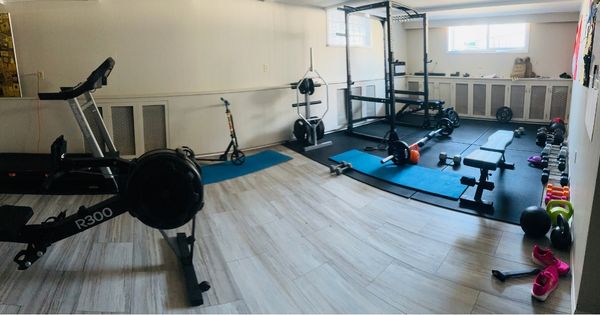 Home gym setup in a bright basement with a rowing machine, squat rack, weight bench, dumbbells, kettlebells, and exercise mats arranged neatly on light wood flooring.